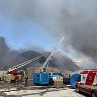 Black smoke surrounds a damaged warehouse, with emergency vehicles attending