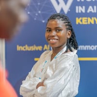 A smiling young woman with crossed arms stands in front of a conference poster for an event on women in AI in Kenya