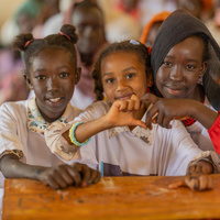 Three Sudanese refugee pupils sit at a desk, smiling, two of them holding their hands together to make a heart shape.