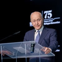 A man in a suit delivers a speech standing at a clear lectern in front of a dark background.