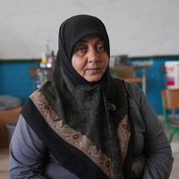 A woman in a headscarf sits in a school classroom with household items on desks in the background