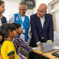 Barham Salih and UNHCR colleagues speak with a family sitting behind a desk with a computer. Everyone is smiling.
