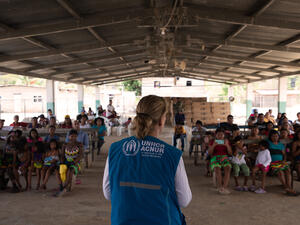 A woman in a branded UNHCR vest addresses a group of people in an open-sided building.