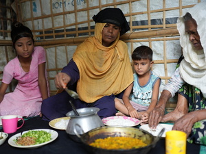 A mother and a grandmother eat a meal sitting on the floor of their shelter with two young children.