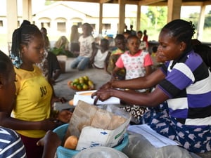 At a market, a woman hands over a small baguette wrapped in paper to a customer.