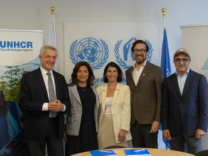 Five people smile and pose for the camera behind a table, with UNHCR banners in the background.