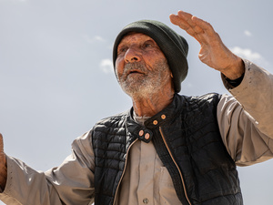 An elderly man wearing a cap and holding a walking stick stands outside beneath a clear sky with his hands raised