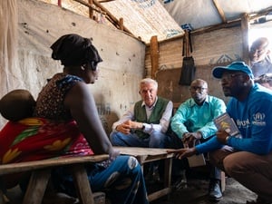 A woman with a young child strapped to her back sits opposite three men inside a tented structure