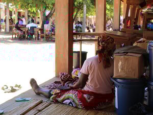A woman sits in the shade on the wooden terrace of a school, leaning back against boxes of aid supplies 