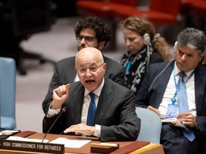 A man gestures while speaking behind a desk with people sitting in the background