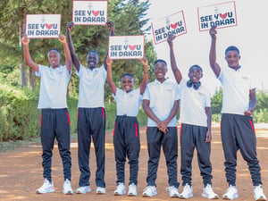 Six smiling young athletes wearing training shoes and clothes hold up signs reading 'see you in Dakar' on a wide dusty road 