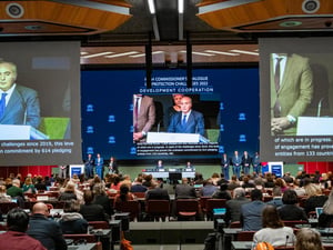 Three large screens show a projection of a speaker in a huge conference room.