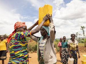 Dans un paysage aride, des femmes aux vêtements colorés collectent de l'eau dans des bidons qu'elles portent sur leur tête ou dans leurs mains.