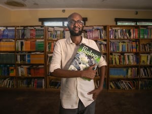 Abdullahi Mire stands in a library in front of a huge bookshelf, clutching a biology book
