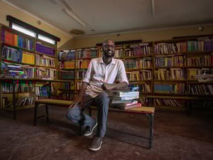 A man sits in a library, surrounded by books and leaning on a pile of books