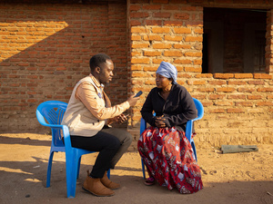 A man interviews a woman outside on two plastic chairs, in front of a brick building