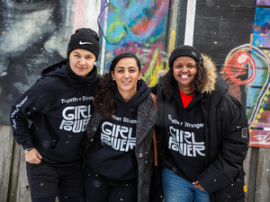 Three women wearing 'Girl Power' jumpers smile at the camera, their arms around each other, in front of a colorful building