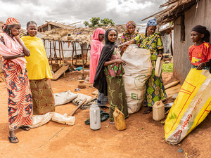 A group of women in colourful dresses stand outside a mud wall hut as they fill two large sacks with belongings