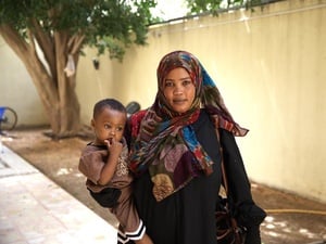 A woman wearing a headscarf stands in a walled courtyard holding a toddler.