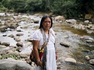 Une femme portant une longue robe blanche et un sac tissé coloré sur l'épaule se tient sur la rive d'une rivière rocheuse peu profonde.