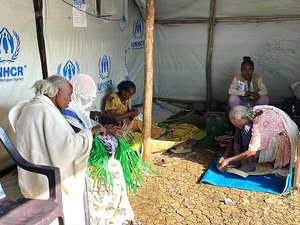 Several women sit inside a UNHCR tent and craft woven items