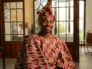 A smiling woman in a patterned traditional dress and headscarft sits in an office