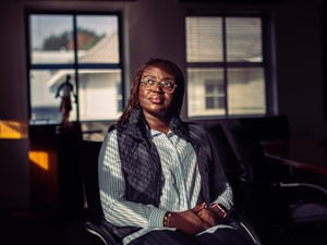 A woman sitting indoors in dappled light, with windows behind her, gazes into the distance