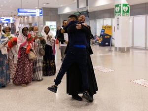 A group of family members embrace tightly at the Athens International Airport, while others stand nearby, smiling and clapping