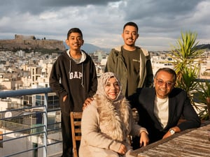 A middle-aged couple sit on a balcony with their two sons standing behind them, with a view of Athens behind them. 