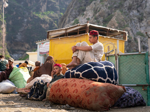 Two brothers wait at a bus stop in front of the mountains, sitting on large piles of belongings.