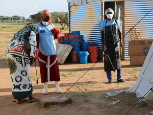 Health workers wearing face masks are pictured outdoors with disinfection equipment. They are standing in front of a metal shed and piles of buckets.
