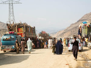 Afghan returnees walk along a dirt road beside loaded trucks.