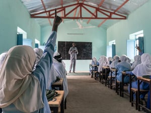 A rear view of a pupil raising her hand at the back of a classroom filled with schoolgirls in uniform sitting at rows of desks 