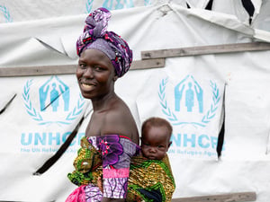 A smiling woman carries her baby boy on her back in front of a UNHCR tent.