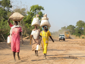 Three friends, Sudanese refugee women, walk together along a dirt road, bags balanced on their heads, smiling and laughing.