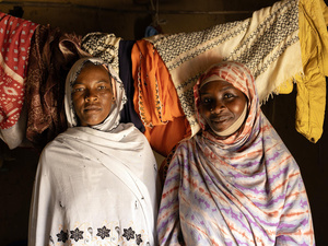 Two women stand next to each other inside a room with washing hanging on a line behind them