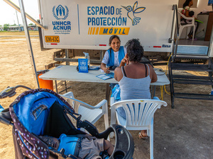 A female member of UNHCR staff speaks to a woman across a table with a baby stroller in the foreground and a truck in the background
