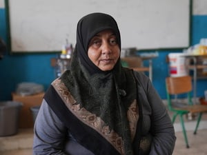 A woman in a headscarf sits in a school classroom with household items on desks in the background
