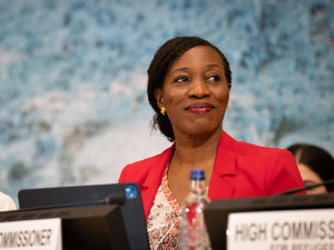 A smiling woman wearing a red jacket sits on a stage behind a microphone 