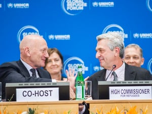 Two men shake hands on a podium behind the signs 'Co-host' and 'High Commissioner for refugees'.