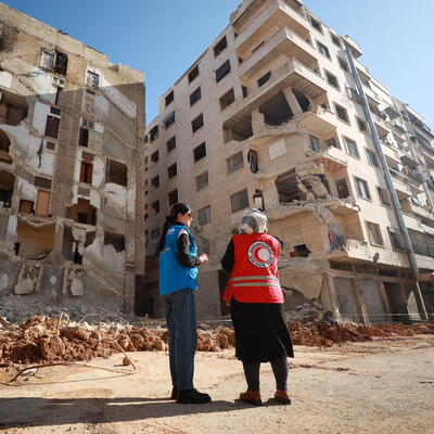 Une employée du HCR et une employée de la Croix-Rouge sont debout devant un bâtiment détruit.