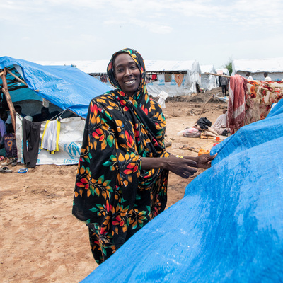 A woman smiles as she pulls a blue tarpaulin into place over a makeshift shelter.