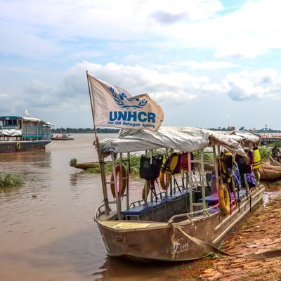 Wooden boats float on a wide, muddy river. One bears a UNHCR flag.