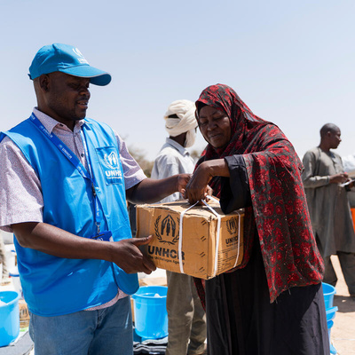 A man wearing blue UNHCR cap and waistcoat hands a woman a cardboard package 