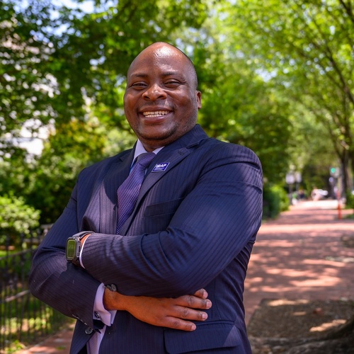 Bahati stands in a park in a suit, crossing his arms and smiling broadly