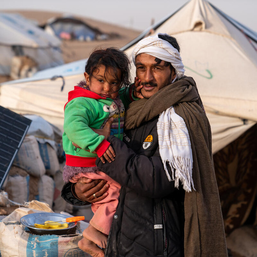 An internally displaced man in Afghanistan, wrapped in blankets, holds a small child. Behind him is a refugee tent settlement.