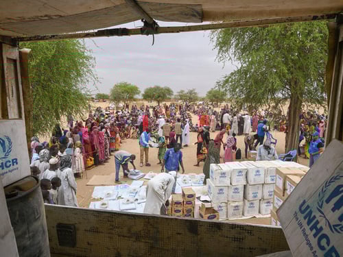 A view from the back of a truck showing workers unloading relief supplies for refugees waiting nearby.