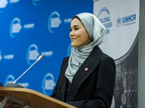 A woman speaks into a microphone at a podium in front of a UNHCR/Global Refugee Forum banner.