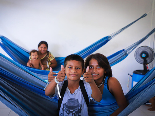 Two children sit in a hammock giving a thumbs up, with a mother and baby in the hammock behind them