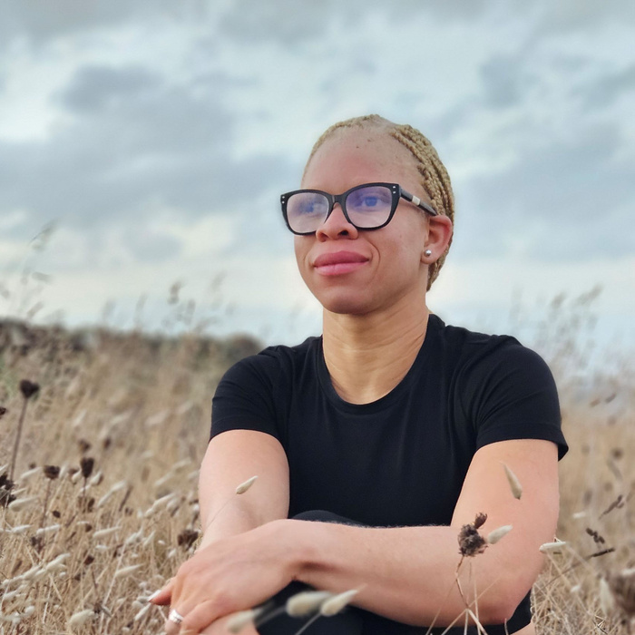 An Albino woman wearing glasses sits in a field looking into the distance, with her hair tied back
